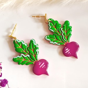 Op de afbeelding: Een paar grillige oorbellen met een bietenontwerp. Elke oorbel heeft een levendige magenta biet met groene bladeren omlijnd in wit. De oorbellen hebben goudkleurige pinnen voor doorboorde oren.