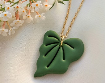 May include: A green leaf pendant necklace with a gold chain. The leaf has a matte finish and detailed veining. The necklace is displayed on a white surface with white flowers in the background.