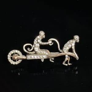May include: A silver brooch with two monkeys, one riding a wheelbarrow, the other pushing it. Both monkeys are covered in sparkling rhinestones.