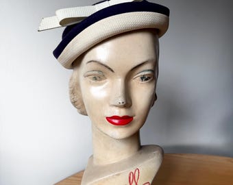 Puede incluir: Una cabeza de maniquí de estilo vintage con rasgos faciales pintados, incluidos labios rojos, que lleva un sombrero azul marino y crema con un lazo prominente. La base del maniquí presenta el texto en letra roja que dice a Jacoll Hat.