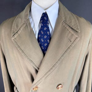 Puede incluir: Un abrigo tipo trench beige de doble botonadura con cinturón a juego, usado sobre una camisa blanca con cuello y una corbata azul con estampado de cachemir. El abrigo tiene botones grandes y un diseño clásico.