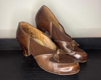 Op de afbeelding: Een paar vintage bruin leren schoenen met een strikdetail op de wreef. De schoenen hebben een ronde neus en een kleine hak. De bovenkant heeft een geperforeerd ontwerp. De schoenen worden op een zwart oppervlak weergegeven.