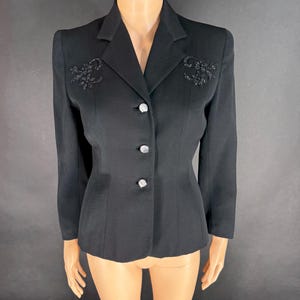 May include: A black blazer featuring a notched lapel, long sleeves, and three silver-toned buttons down the front. The upper chest area on both sides is decorated with black beaded floral embroidery. The garment is displayed on a mannequin.