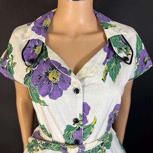 May include: A vintage white dress with a floral pattern of large purple flowers, green leaves, and yellow accents. The dress has a collared neckline, short sleeves, and a matching belt. Black buttons fasten down the front.