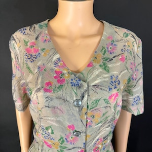 Puede incluir: Un vestido vintage con estampado floral. El vestido presenta un escote en V, mangas cortas y una botonadura frontal. La tela tiene un fondo verde claro con acentos florales rosas, azules y amarillos. El vestido se exhibe en un maniquí.