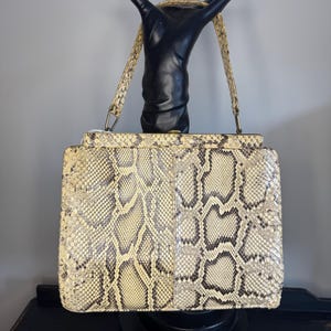 以下が含まれることがあります： クリーム、タン、黒色のスネークスキン柄のヴィンテージハンドバッグ。長方形のバッグは、クラスプクロージャーとマッチするスネークスキンのストラップを備えた構造的な形状をしています。バッグは黒い手型のマネキンに表示されています。