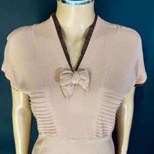 Peut inclure: Robe rose pâle de style vintage, à manches courtes et col en V. L'encolure et les bords des manches sont bordés d'un tissu marron foncé. Un petit nœud est centré à l'encolure, et le corsage présente des plis horizontaux.
