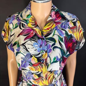 Puede incluir: Vestido vintage con estampado floral con un vibrante patrón de flores moradas, amarillas y rojas sobre un fondo crema. El vestido tiene cuello, mangas cortas y botones en la parte delantera. La falda es plisada.
