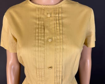 May include: A vintage mustard yellow dress with short sleeves and a pleated bodice. The dress features a round neckline and three buttons down the front. The skirt is pleated, and the dress is displayed on a mannequin.