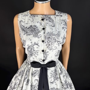 May include: A sleeveless dress featuring a white base with a black paisley and floral print. The bodice includes a square neckline and a vertical row of five black buttons. A solid black ribbon bow is tied at the waist, accentuating the full skirt.