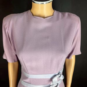 May include: A lavender short-sleeved dress featuring a round neckline and structured shoulders. The waist area is accented with two horizontal light blue bands and two decorative bows tied on the right side, creating a layered visual effect.