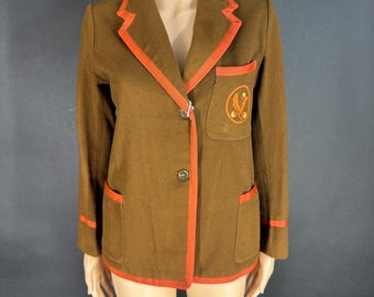 May include: A brown blazer featuring bright orange piping along the lapels, cuffs, and hem. The jacket has two lower pockets, one chest pocket with an embroidered circular emblem, and two front buttons. The design includes long sleeves and a collar.