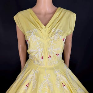 May include: A pale yellow sleeveless dress featuring a V-neckline and a full skirt. The bodice and skirt are decorated with white embroidered leaf patterns and small red circular accents arranged in clusters throughout the garment.