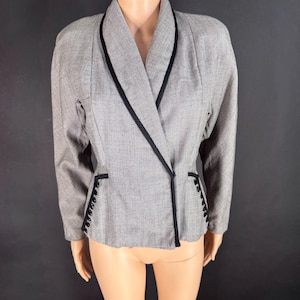 May include: A grey blazer with a fine micro-check pattern, featuring a shawl collar trimmed in black piping. The garment includes long sleeves, structured shoulders, and two decorative side panels, each adorned with a vertical row of small black buttons.