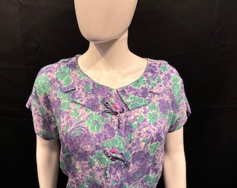 Könnte beinhalten: Ein lila-grünes Blumenkleid mit kurzen Ärmeln und Knopfleiste. Das Kleid hat ein tailliertes Oberteil und einen weiten Rock.