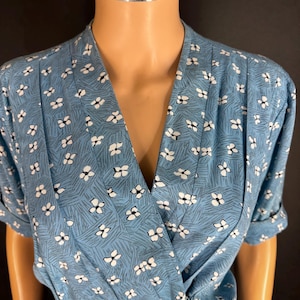 May include: A blue wrap-style blouse featuring a repeating pattern of small white four-petaled flowers and thin dark gray diagonal lines. The garment has a V-neckline, short sleeves with folded cuffs, and dark buttons along the lower front section.