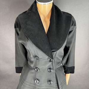 Puede incluir: Una chaqueta negra de doble botonadura con un amplio cuello chal de terciopelo, mangas tres cuartos con puños de terciopelo y ocho botones negros redondos dispuestos en dos filas verticales. La chaqueta tiene costuras horizontales visibles.