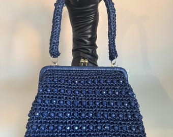 Puede incluir: Un bolso de mano azul marino con cuentas y un asa trenzada. El bolso se exhibe en una mano de maniquí negra. El bolso tiene forma rectangular con cierre de broche y está adornado con pequeñas cuentas azul oscuro.