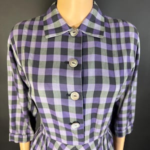 May include: A vintage dress with a purple, black, and grey checked pattern. The dress has a collared neckline, button-down front, and three-quarter sleeves. The skirt is pleated, and the dress is displayed on a mannequin.