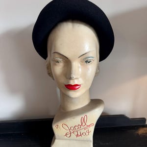 May include: A black felt hat on a white mannequin head. The mannequin head is labeled "a Jacoll Hat Reg'd".