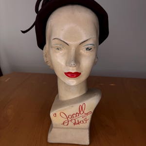 Può includere: Un cappello marrone scuro con un dettaglio a fiocco è esposto su una testa di manichino. Il manichino ha il rossetto rosso e il testo "a Jacoll Hat" in rosso.