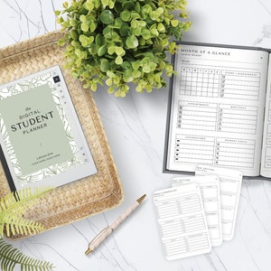 Könnte beinhalten: Ein Flatlay-Bild eines grau-weißen Planers mit dem Text "The Digital Student Planner" auf dem Cover. Der Planer ist auf einer Seite mit einem Monatskalender und einem Abschnitt zum Setzen von Zielen geöffnet. Es gibt auch mehrere andere Planerseiten, einen Stift, eine Tasse Kaffee, eine kleine Holzkiste mit Bleistiften und Pflanzen.