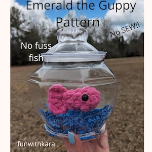 May include: A pink crocheted guppy fish in a clear glass jar with blue shredded paper. The jar has a lid and the text "Emerald the Guppy Pattern No SEW!! No fuss fish funwithkara" is visible on the image.