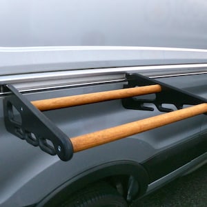 May include: A black metal and wood rack attached to the side of a gray van. The rack has two wooden dowels and is designed to hold items. The van's side has a sliding door track.