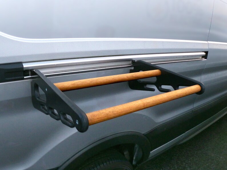 May include: A black metal and wood rack attached to the side of a gray van. The rack has two wooden dowels and is designed to hold items. The van's side has a sliding door track.