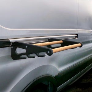 May include: A black and wood-toned van-mounted rack with two horizontal bars and multiple hooks. The rack is attached to the side of a silver van. The background shows a beach scene with palm trees and the ocean.