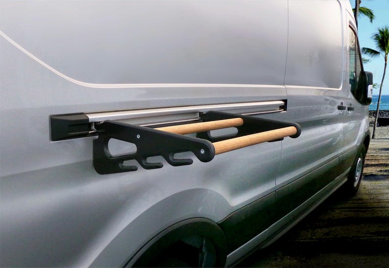 May include: A black and wood-toned van-mounted rack with two horizontal bars and multiple hooks. The rack is attached to the side of a silver van. The background shows a beach scene with palm trees and the ocean.