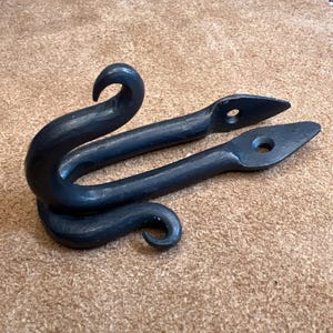 May include: A hand-forged, dark grey metal hook with a curved design. The hook has two pointed ends with drilled holes and a curled top and bottom. The metal has a textured appearance.