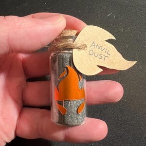 May include: A small glass bottle filled with gray powder, sealed with a cork and twine. An orange flame graphic is on the bottle, and a tag reads "ANVIL DUST". The bottle is held in a hand.