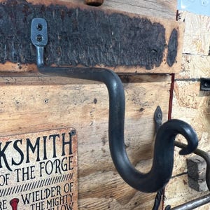 May include: A hand-forged dark gray metal hook with a curved, S-shaped design. The hook features a flat mounting plate secured with two screws to a rough, textured wooden board. The metal surface has a matte finish with visible hammer marks.