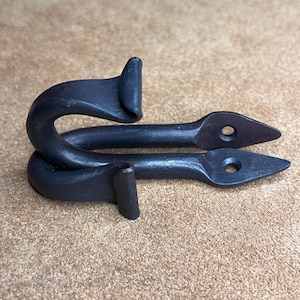 May include: A pair of black, forged iron gate latches. Each latch features a curved hook and a pointed end with a hole for mounting. The metal has a textured, matte finish. The latches are designed to interlock.