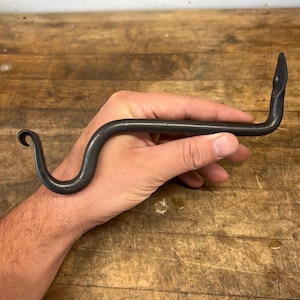 May include: A hand-forged, dark gray metal hook with a snake-like design. The hook curves in a serpentine shape, with a curled end and a pointed tip. The hook is held in a hand against a wooden background.