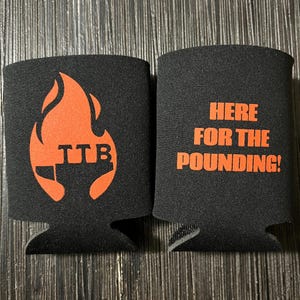 May include: Two black can coolers. One features an orange flame design with the letters "TTB". The other cooler has the phrase "HERE FOR THE POUNDING!" in orange text.