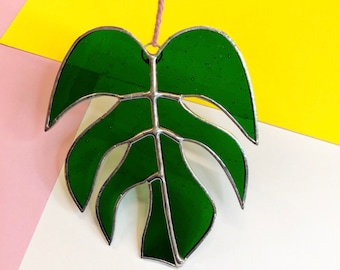 May include: A green stained glass monstera leaf hanging from a pink string. The leaf is detailed with silver metal framing.