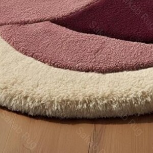 May include: Close-up of a round rug with a layered design. The rug features concentric arcs of color, including cream, light pink, and a deeper rose hue. The rug has a soft, textured appearance, suggesting a plush material.