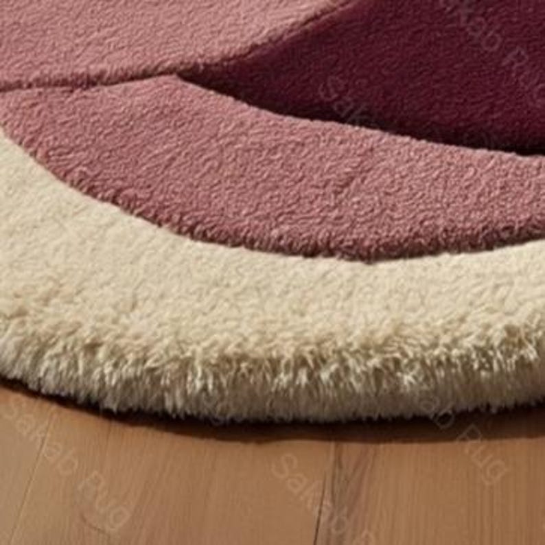 May include: Close-up of a round rug with a layered design. The rug features concentric arcs of color, including cream, light pink, and a deeper rose hue. The rug has a soft, textured appearance, suggesting a plush material.