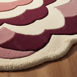 May include: A floral-shaped rug in shades of pink, cream, and burgundy. The rug has a soft, textured surface with a layered design. The rug's edges are rounded, and it sits on a wooden floor.