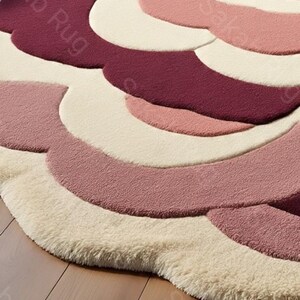 May include: A round rug with a floral design in shades of pink, cream, and burgundy. The rug has a soft, textured appearance, with a layered petal-like pattern. The rug is placed on a wooden floor.