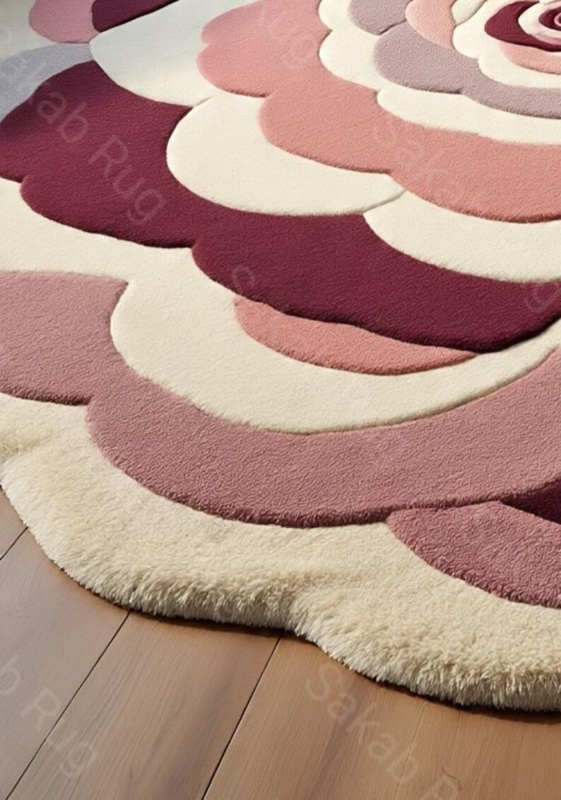 May include: A round rug with a floral design in shades of pink, cream, and burgundy. The rug has a soft, textured appearance, with a layered petal-like pattern. The rug is placed on a wooden floor.