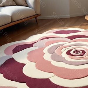 May include: A large, round rug with a floral design. The rug features a layered pattern of petals in shades of pink, white, and burgundy. The rug is placed on a hardwood floor, with a sofa visible in the background.