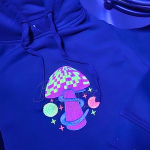 May include: Black hoodie featuring a neon green, pink and yellow mushroom graphic. The mushroom has a checkered pattern on the top. The hoodie has the text "Bloo Moaz atelier" in white on the bottom right corner.