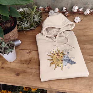May include: A beige hoodie with a sun and moon design embroidered on the front. The design features a sun with a face and a crescent moon with a cloud. The text "El Sol y La Paz" is embroidered above the design.