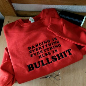 May include: A bright red crewneck sweatshirt featuring black embroidered text that reads DANCING IS EVERYTHING EVERYTHING ELSE IS BULLSHIT. The garment has long sleeves and a ribbed neckline, accompanied by a spool of black thread and small scissors.