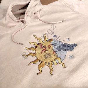 May include: A cream-colored hoodie with an embroidered design of a sun and moon with stars. The sun is yellow with a red face and the moon is gray with a cloud.