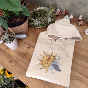 May include: A cream-colored hoodie with an embroidered design of a sun and moon with a face. The sun is yellow and the moon is gray. The text "RooBaz" is embroidered on the hoodie.