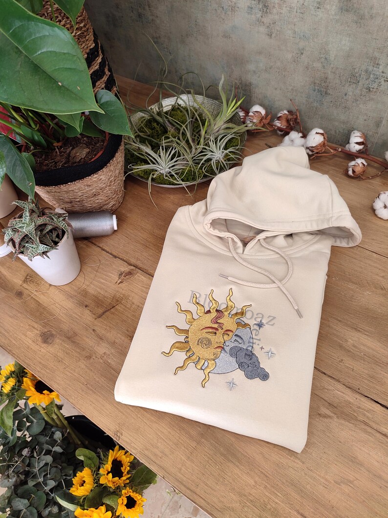 May include: A cream-colored hoodie with an embroidered design of a sun and moon with a face. The sun is yellow and the moon is gray. The text "RooBaz" is embroidered on the hoodie.